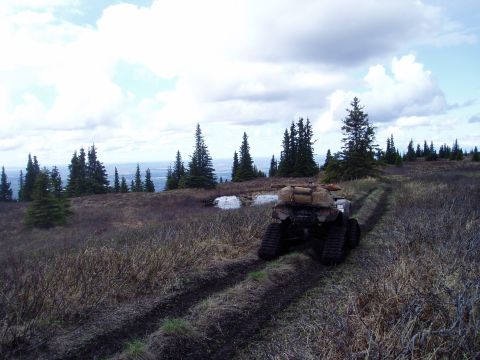 2000 Ford E350 - An all-terrain vehicle with tracks and cargo navigates a muddy trail through a wooded, hilly landscape under a cloudy sky.