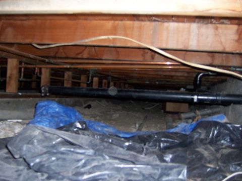 1991 Ford Bronco - Underneath a structure, a crawl space contains plumbing, wiring, and debris covered by plastic sheeting.