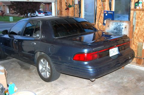 1985 Mercury Grand Marquis - Rear view of a dark blue sedan with tinted windows and a spoiler, parked inside a garage.