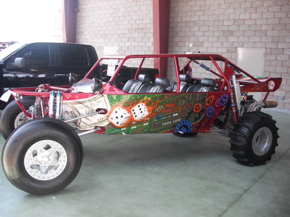 Red dune buggy with dice and dollar bill graphics, parked next to a black pickup truck