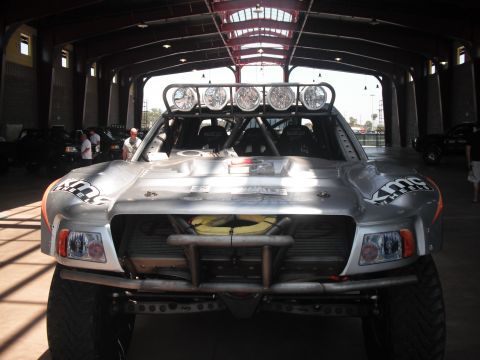 1986 Ford Bronco - Silver off-road race truck with a roll cage and large light bar parked indoors.
