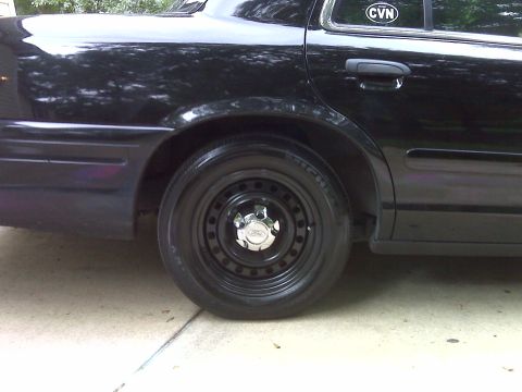 2001 Ford Crown Victoria - Close-up of a black sedan's rear wheel, showing a steel rim and a chrome hubcap. A 'CVN' sticker is visible on the window.