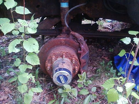 1990 Ford Bronco - This rusty wheel hub and suspension component are overgrown with weeds, suggesting the vehicle is in disrepair.