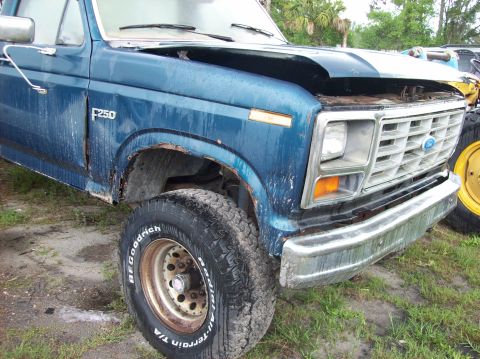 1985 Ford F-250 - This blue Ford F-250 truck shows significant rust and damage, with its hood partially open.