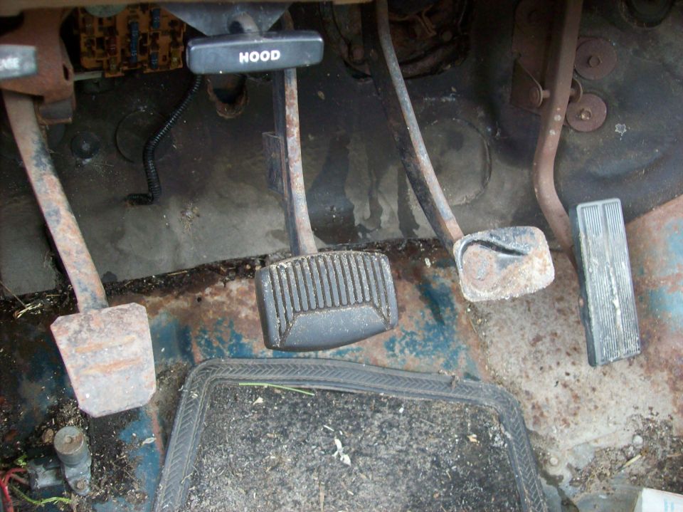 Floor pedals and hood release lever in a vehicle interior. The floor is rusty and dirty