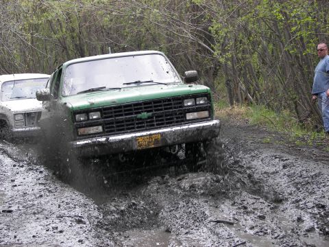 1990 Chevrolet Suburban - Two trucks are navigating a muddy trail with trees on either side.