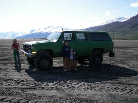1990 Chevrolet Suburban - A family poses with their lifted green SUV in a vast, sandy landscape with snow-capped mountains.