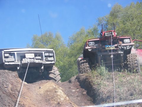 1990 Chevrolet Suburban - Two heavily modified off-road vehicles navigate a challenging dirt terrain under a clear blue sky.