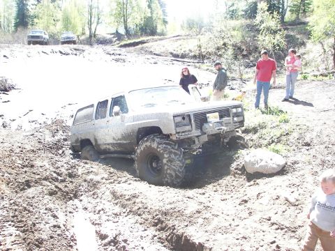1990 Chevrolet Suburban - This lifted SUV is deep in the mud, with onlookers observing the off-road challenge.