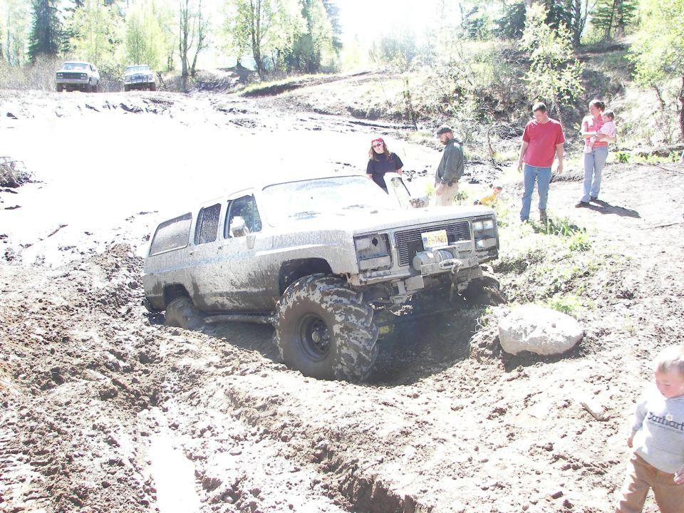 Muddy SUV stuck in a rut with people watching. Two other vehicles are visible further up the hill