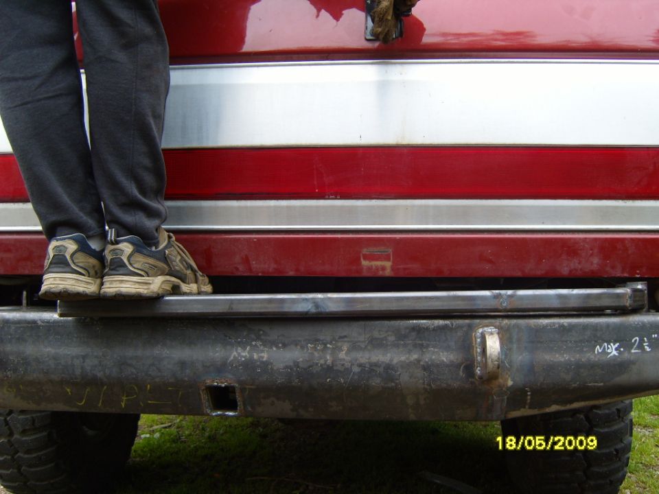 Close-up of a black aftermarket rear bumper with a tow hitch receiver and a metal step. Red and silver stripes are visible above the bumper