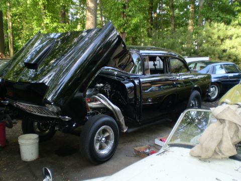 1955 Chevrolet Bel Air - A black 1955 Chevrolet Bel Air is shown with its hood open, revealing the engine and custom exhaust. Another vehicle is visible in the back…