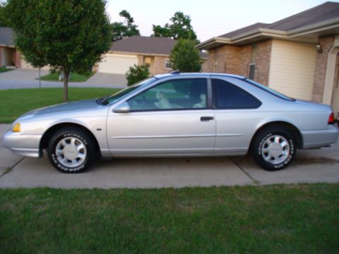 1995 Ford Thunderbird - A silver coupe is parked on a concrete driveway in front of a residential house.