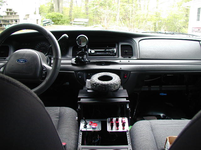 Car interior showing dashboard, steering wheel with Ford logo, center console with switches, and a spare tire on top