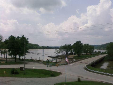 1985 Ford LTD Crown Victoria - A bridge crosses a wide river, with RVs and trees lining the banks under a cloudy sky.