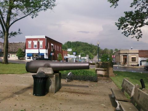 1985 Ford LTD Crown Victoria - A historic cannon is prominently displayed in the center of a town square, surrounded by buildings and greenery.