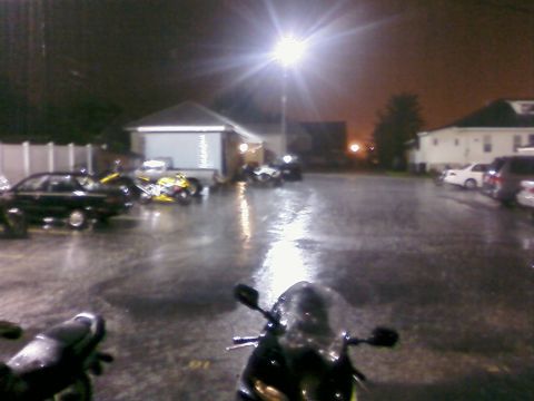 1985 Ford LTD Crown Victoria - A motorcycle's windshield is visible in the foreground of a wet parking lot at night, with several other vehicles parked around.