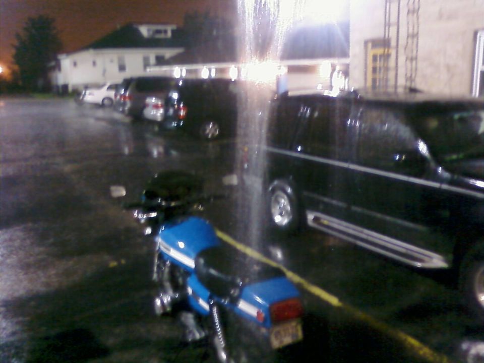 Blue motorcycle with white stripe parked on wet asphalt at night. Black van and other vehicles in background
