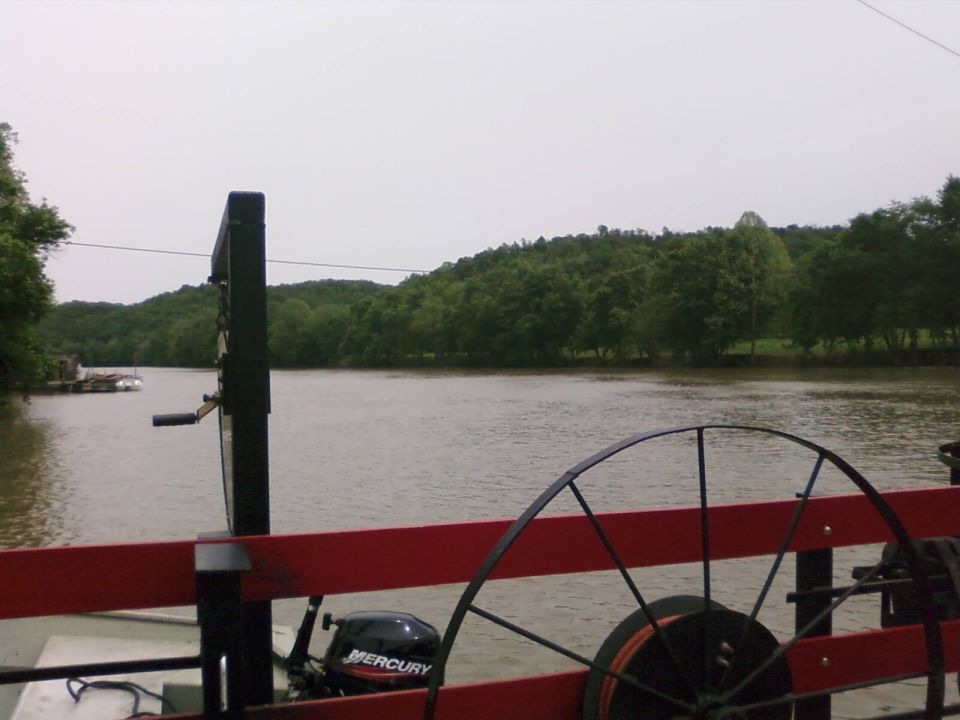 View from a boat looking out at a wide river with tree-covered hills in the background. A Mercury outboard motor is visible on the right