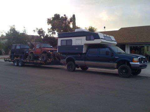 1987 Suzuki Samurai - A dark blue pickup truck tows a trailer carrying two off-road vehicles, including a red Suzuki Samurai.