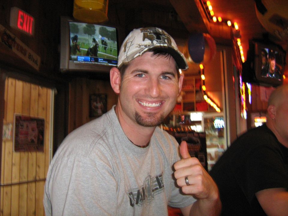 Man in a camouflage hat and grey t-shirt gives a thumbs up in a bar with TVs showing horse racing