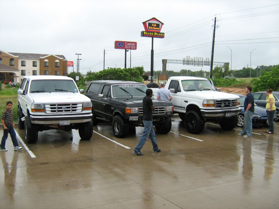 Three lifted Ford trucks and SUVs parked in a lot. The white one on the left looks like a 1992 Ford Bronco, the black one in the middle is a Bronco, and the white one on the right is a Ford F-150