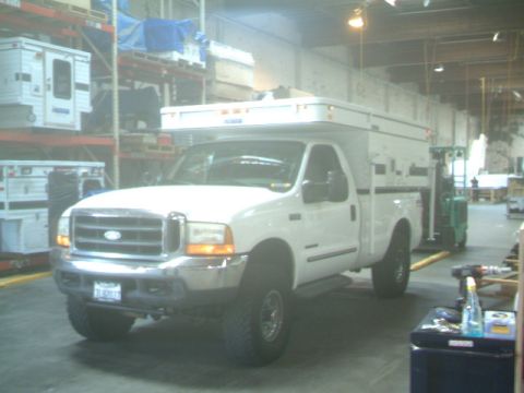 2000 Ford F-250 - A white Ford pickup truck equipped with a camper shell is parked inside a warehouse, with storage shelves in the background.