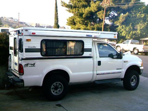 2000 Ford F-250 - A white Ford F-250 pickup truck is equipped with a Grandby camper shell, seen from the side with another white pickup truck in the backgrou…