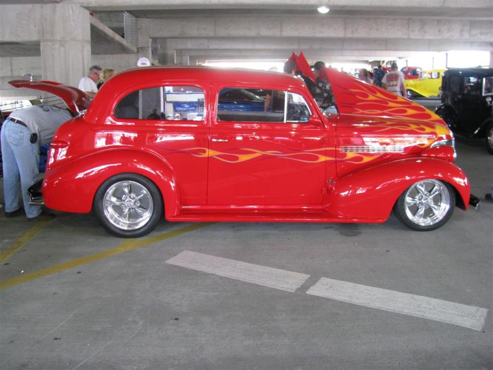 Red hot rod with flame decals and custom wheels, hood open. Another car is visible behind it