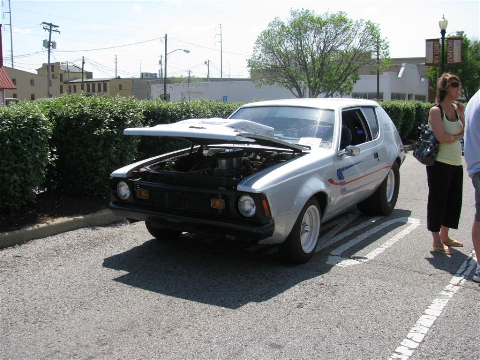 Silver AMC Gremlin with hood open, showing the engine. Two people stand nearby