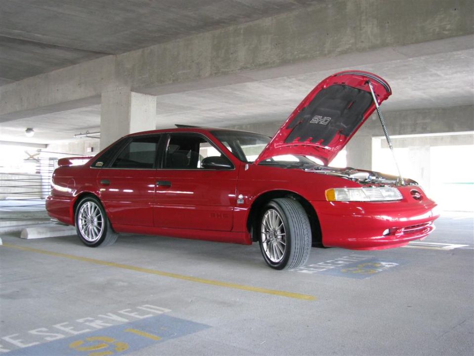 Red sedan with hood open parked in a garage. Has a spoiler and aftermarket wheels