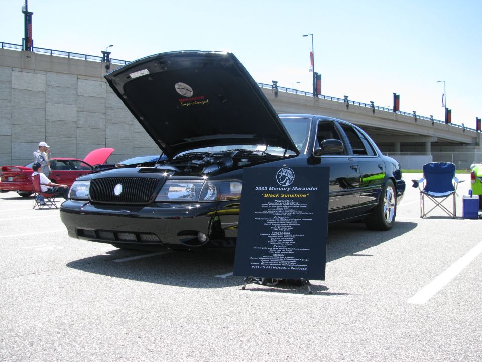 Black 2003 Mercury Marauder with hood open, parked at a car show. A sign details the car's features
