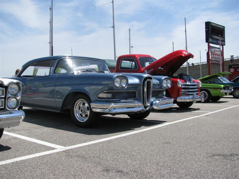 Several classic cars parked in a lot, including a blue sedan and a red pickup truck with its hood open