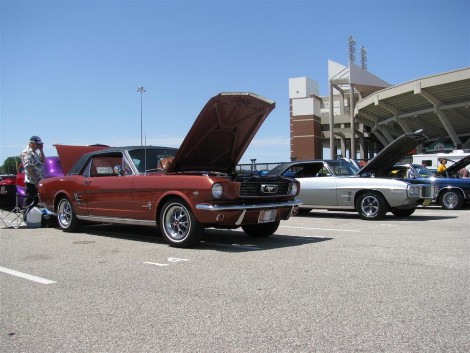 Red 1965 Ford Mustang coupe with hood open, parked next to other classic cars at a show