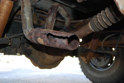 1994 Ford Bronco - Underbody shot of a badly rusted exhaust tailpipe that has rotted through, splitting open along the top near its outlet. Behind it sit a co…