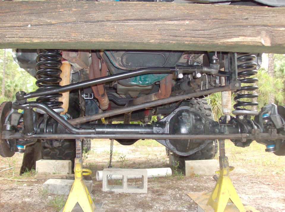 Underside view of a lifted off-road vehicle's front axle, steering linkage, and suspension components, supported by jack stands