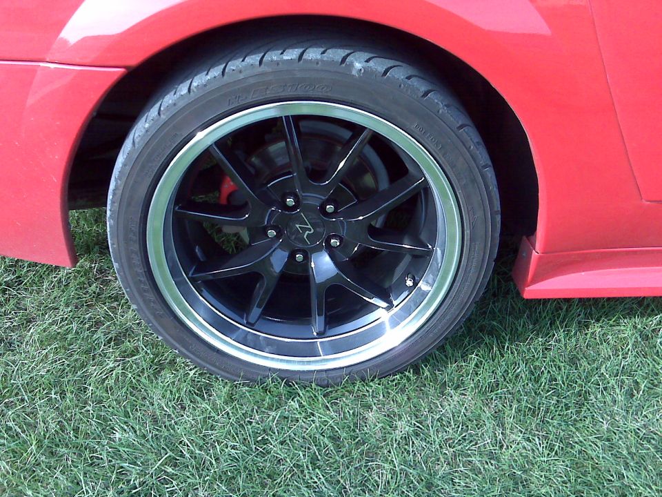 Red 2004 Ford Mustang wheel with black multi-spoke rim and silver lip, parked on grass