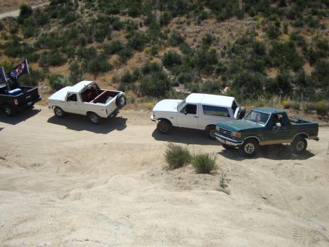 1986 Ford Bronco - Several off-road vehicles are parked on a dusty trail with hills in the background.