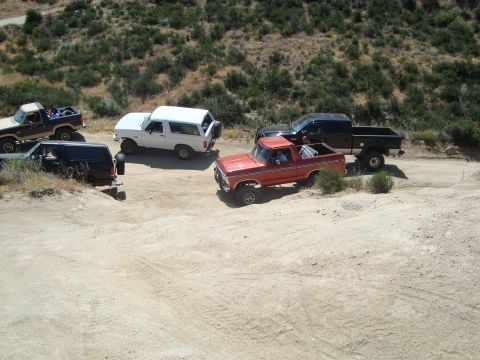 1986 Ford Bronco - A group of off-road vehicles are gathered on a dusty trail in a hilly, arid environment.