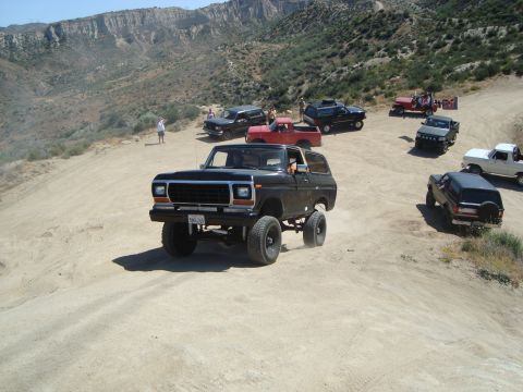 1986 Ford Bronco - Several off-road vehicles are gathered on a dusty trail with mountains in the background.