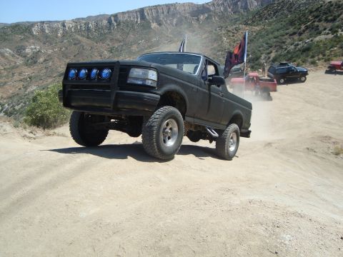 1986 Ford Bronco - A lifted black 1992 Ford F-150 navigates a dusty off-road trail, with other vehicles following in the background.