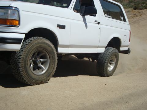 1986 Ford Bronco - White Ford Bronco with off-road tires kicks up dust on a dirt road.