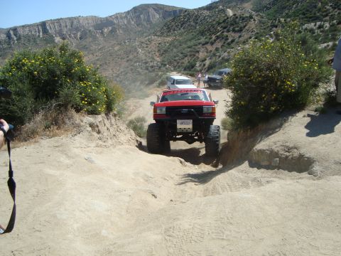 1986 Ford Bronco - A red off-road SUV navigates a dusty trail with other vehicles and people in the distance.