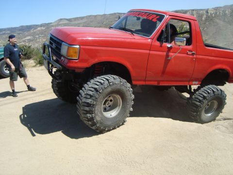 1986 Ford Bronco - Modified red Ford Bronco with oversized tires sits on a dirt road under a clear sky.