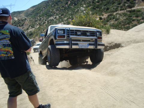 1986 Ford Bronco - A lifted Ford Bronco navigates a sandy off-road path with a person watching.