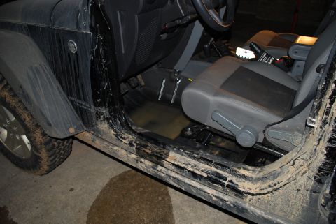 2008 Jeep Wrangler - Doors-off view into the driver's side of a mud-caked black Jeep Wrangler, showing muddy water pooled in the rubber floor pan, cloth bucket…