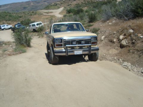 1986 Ford Bronco - A 1986 Ford Bronco navigates an off-road trail, with other SUVs and trucks visible in the distance.