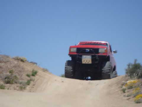1986 Ford Bronco - A red off-road truck navigates a steep dirt incline on a sunny day.