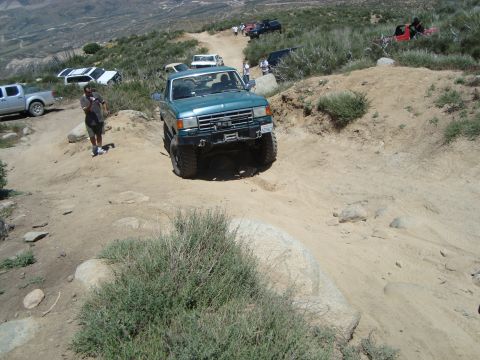 1986 Ford Bronco - A teal Ford F-Series truck navigates a challenging off-road trail with other vehicles present.