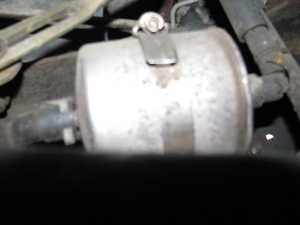 Blurry underbody shot of a silver inline fuel filter with metal band clamp on a 1986 Ford Bronco